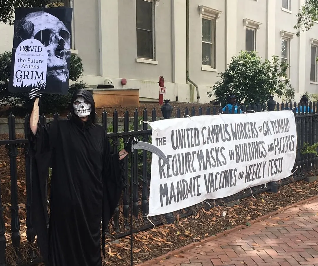 UCW member dressed as Grim Reaper stands by UCW sign: "The United Campus Workers of GA demand—Require masks in buildings and facilities—Mandate Vaccines or Weekly Tests"